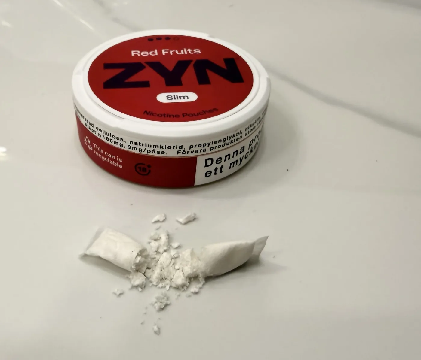 Một hộp ZYN Red Fruits Slim bên cạnh một túi đã bị xé, bột trắng bên trong đổ ra mặt bàn sáng màu.