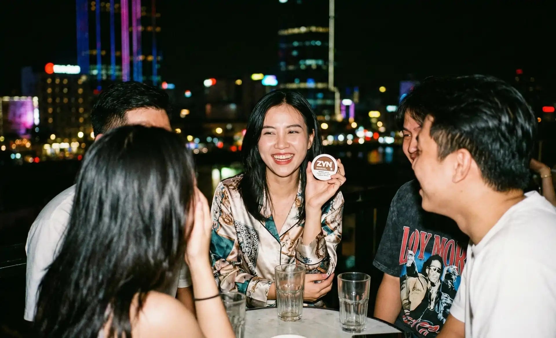 Vietnam nightlife scene with friends laughing at an outdoor rooftop bar, woman showcasing ZYN nicotine pouches against vibrant city lights.